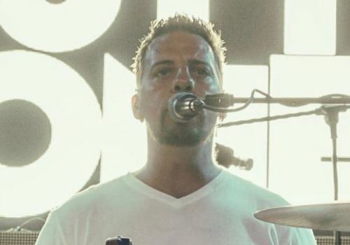 A man in a white shirt is playing the drums and singing into a microphone on stage.