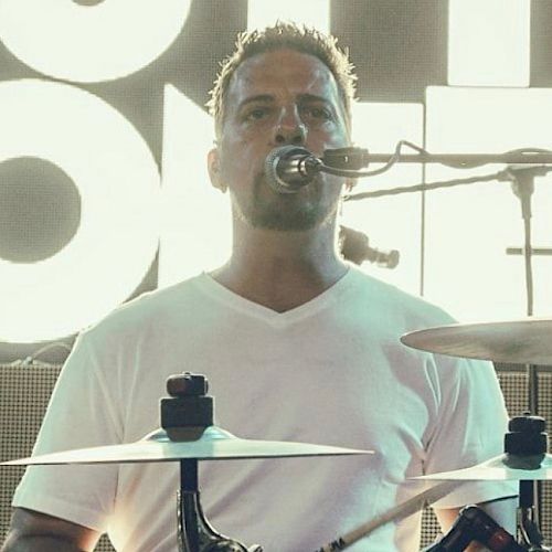 A man in a white shirt is playing the drums and singing into a microphone on stage.