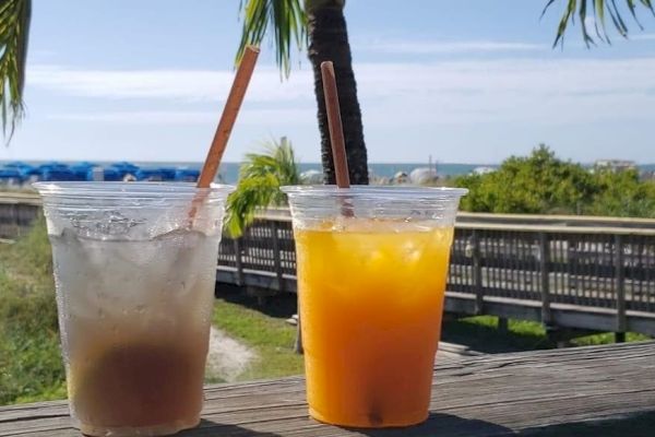 Two drinks in plastic cups with straws are placed on a wooden railing by a beach with a boardwalk, lush greenery, and palm trees in the background.