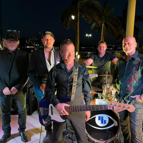 A group of five men, likely a band, pose with musical instruments outdoors at night; drums, bass, and electric guitar are visible.