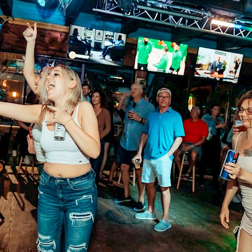 A group of people enjoying a lively bar or club, with one person taking a selfie and others watching televisions and drinking.