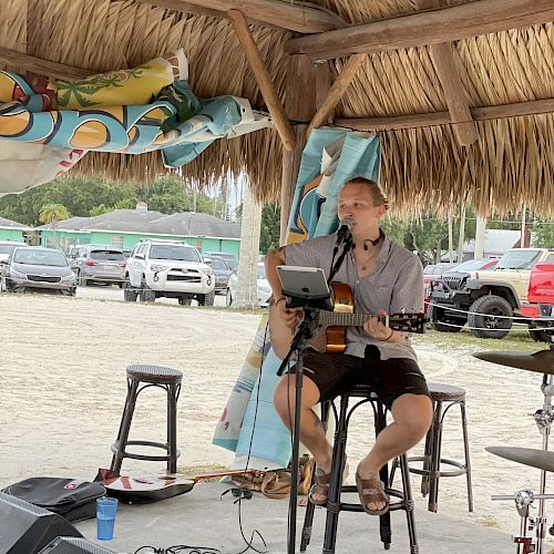 A person is playing guitar and singing into a microphone under a thatched roof, with music equipment around and a parking area in the background.