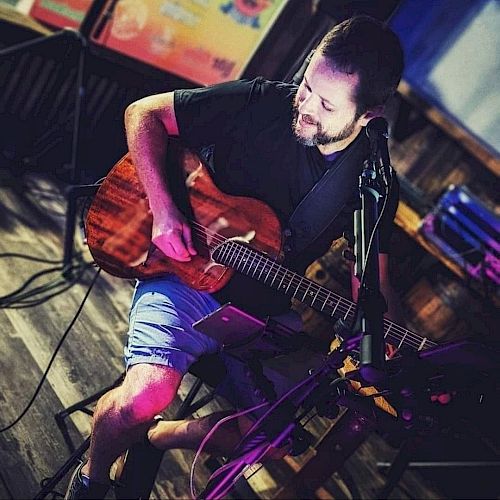 A man is sitting and playing a guitar in a dimly lit setting. There's musical equipment around, and he appears to be performing.