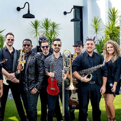 A group of people, each holding musical instruments like a violin, trumpet, ukulele, trombone, saxophone, and a microphone, posing outdoors.