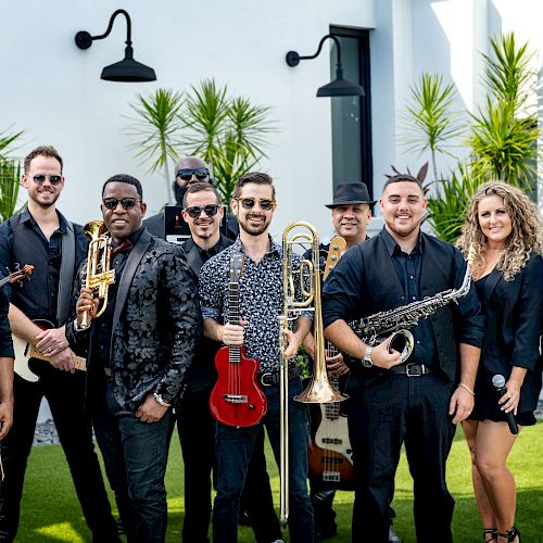 A group of musicians holding various instruments, including a violin, trumpet, trombone, saxophone, guitar, and microphone, outdoors.