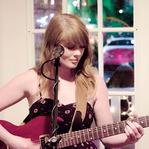 A person with long hair plays an electric guitar and sings into a microphone, standing indoors in front of a window with colorful lights outside the window.