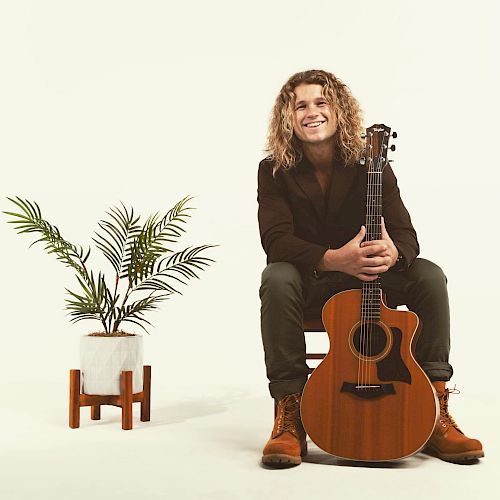 A person with long curly hair is sitting on a chair, holding an acoustic guitar. A plant in a white pot on a wooden stand is beside them.