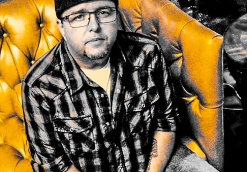 A man wearing a cap and plaid shirt sits on a tufted leather couch next to a table with a glass on it. Text below reads 
