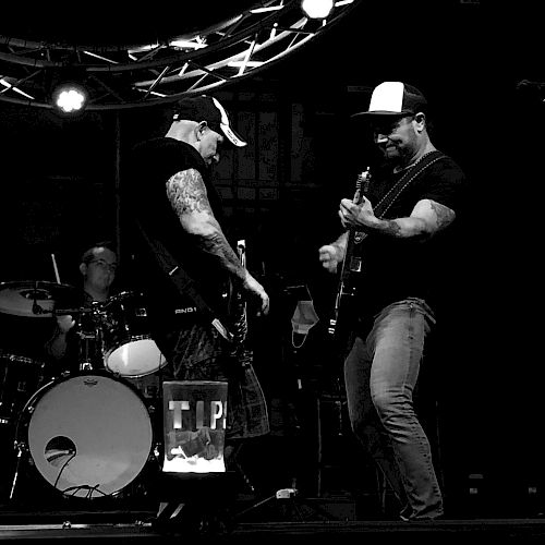 A black and white photo shows three musicians playing on stage: two guitarists in the foreground and a drummer in the background under stage lights.