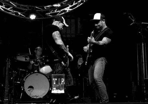 A black and white photo shows three musicians playing on stage: two guitarists in the foreground and a drummer in the background under stage lights.