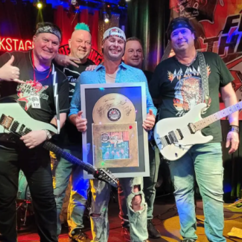 A group of five people with guitars stand on a stage, one holding a plaque, all smiling. There's a drum kit and a banner reading 