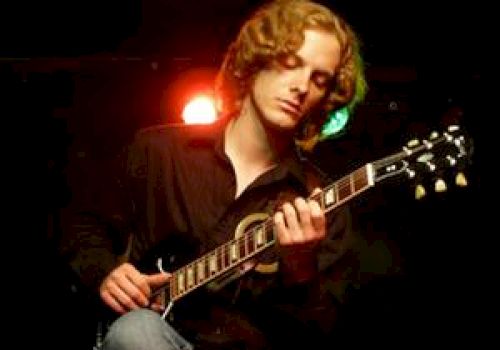 A person with curly hair is seated on a stool, playing an electric guitar under stage lighting.